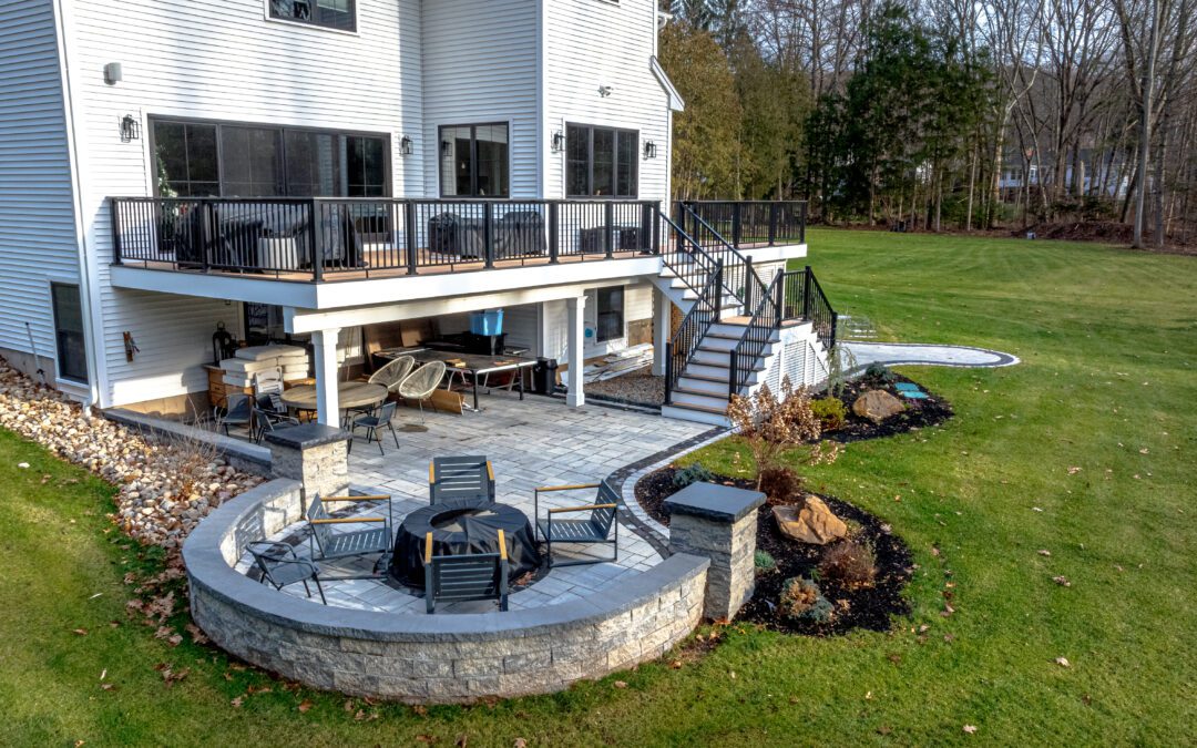 Side angle view highlighting an inviting stone patio with a semi-circular stone seating wall, integrated fire pit, and stylish landscaping beneath an elevated deck attached to a modern home.