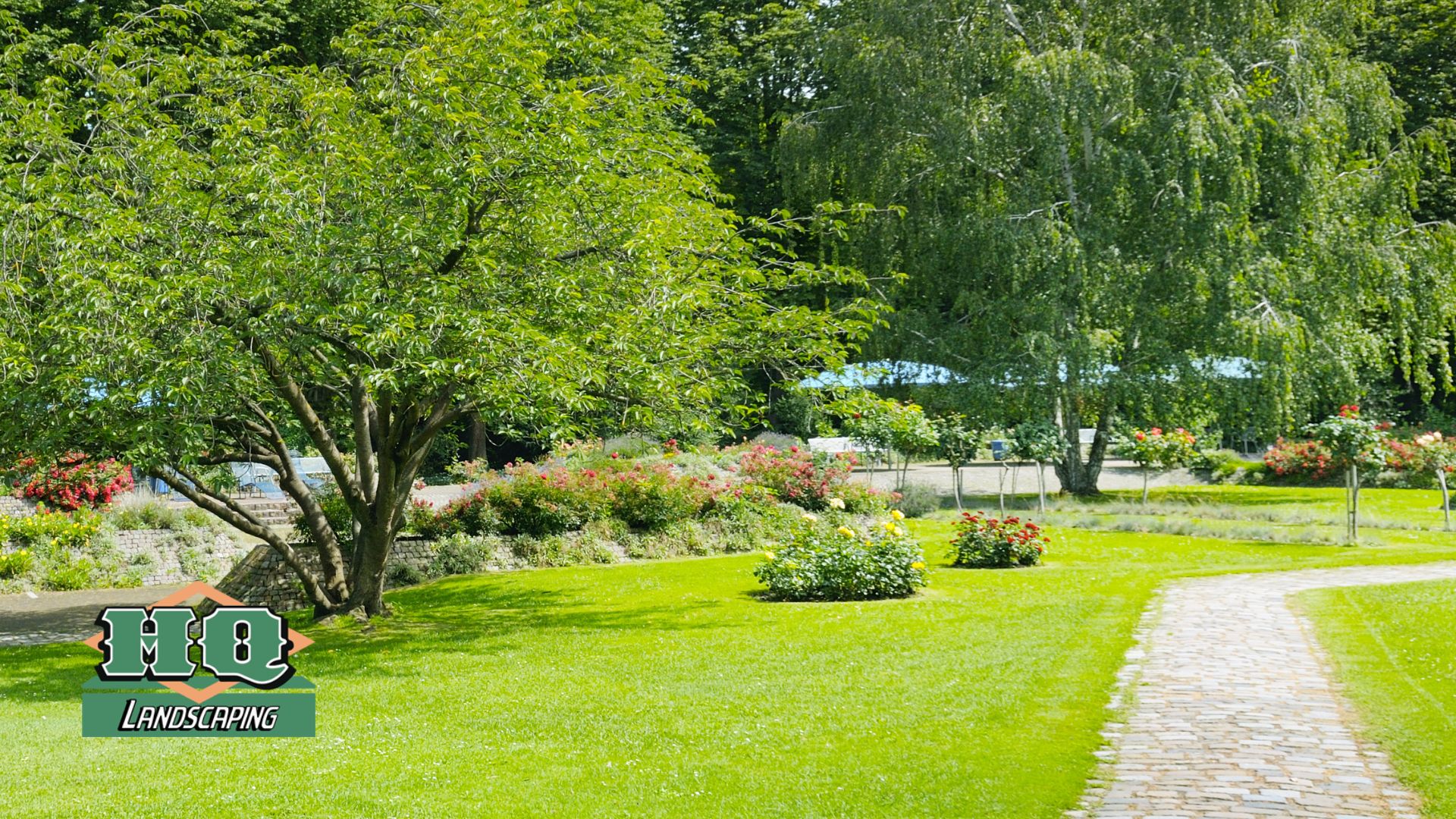 A beautifully landscaped park-like setting with a lush green lawn, a curved stone walkway, mature trees, and colorful flower beds, showcasing HQ Landscaping's professional lawn installation and landscaping services.