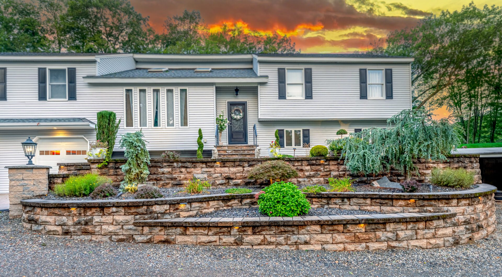 Multi-tier stone retaining wall with landscape lighting and planted shrubs in front of a two-story home in Berlin CT at sunset.